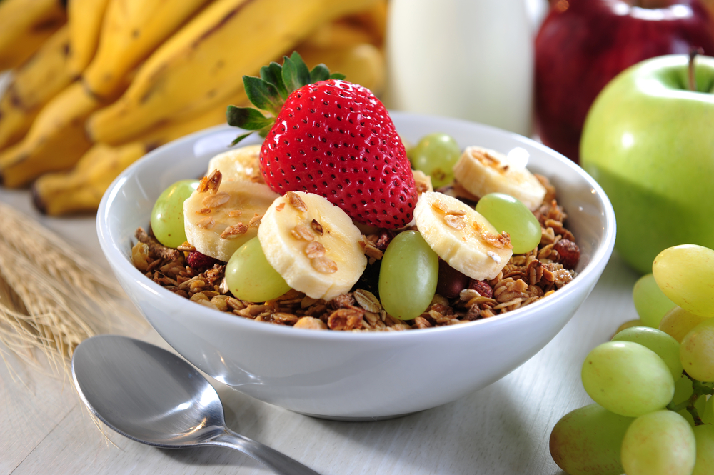 bowl of granola topped with grapes, sliced bananas, and a strawberry next to apples