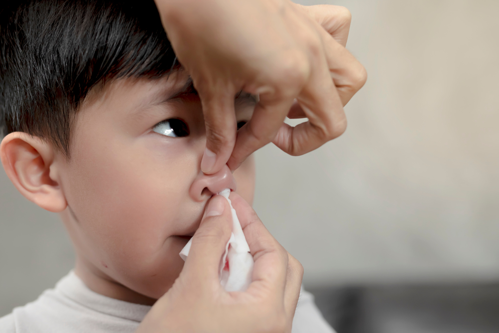 hands applying pressure to little boy's nose while using tissue to soak up blood from nosebleed